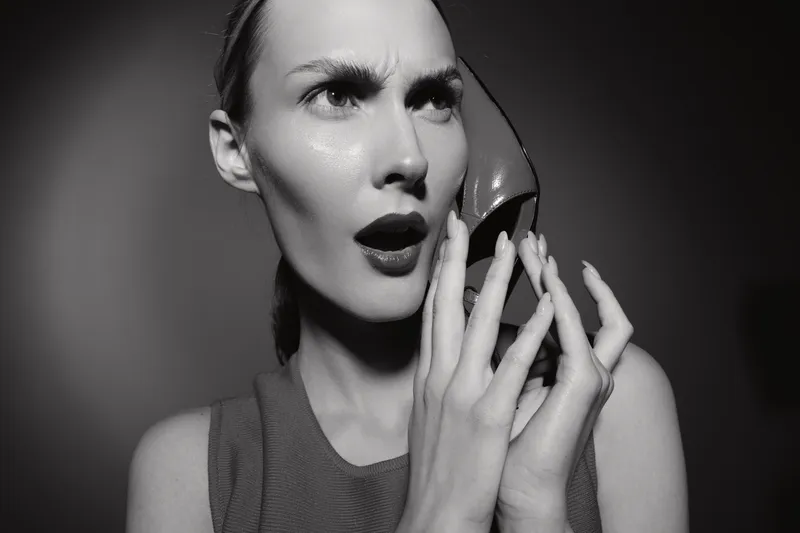 White woman with pale skin and red lips holds red high heel shoe near face with surprised expression. Studio portrait in black and white with dramatic lighting and fashion concept.