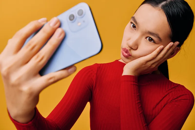 Young woman with dark hair in red sweater takes selfie with light blue smartphone, posing and making playful face against yellow background in closeup portrait photography style.