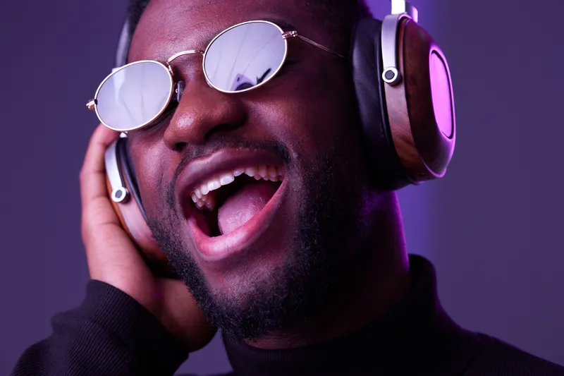 A joyful Black man wearing stylish headphones and sunglasses, passionately singing against a colorful purple and pink background, embodying music and happiness
