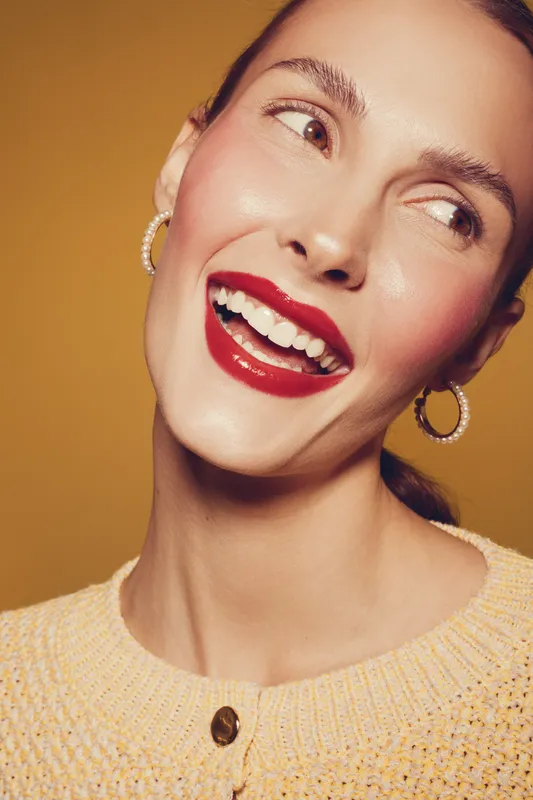 Close-up of smiling woman with red lipstick and hoop earrings wearing yellow sweater on orange background. Concept of beauty, makeup, happiness, and fashion portrait.
