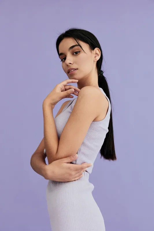 young woman with long dark hair wearing white sleeveless dress posing against pastel purple background, looking confident and serene