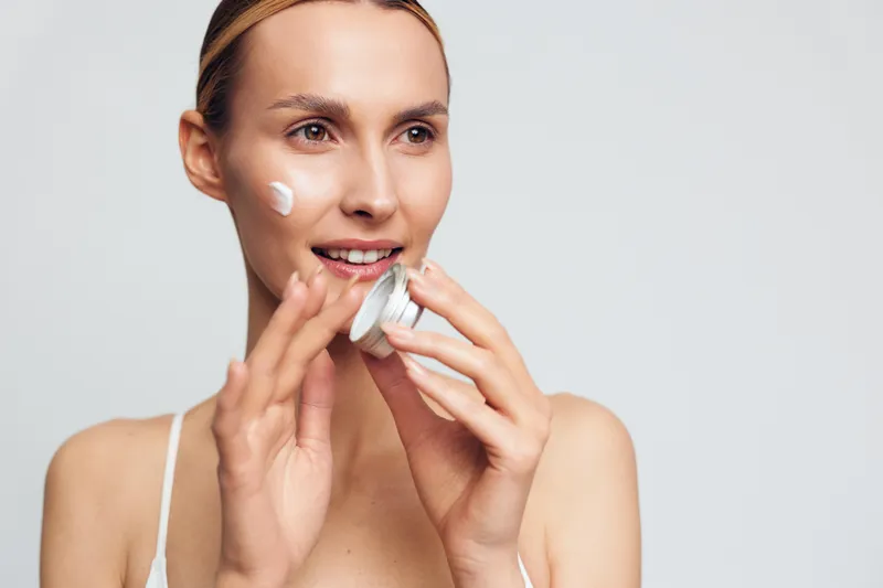 Young woman applying face cream with soft smile, clean skin, natural makeup, skincare routine, bright studio background, healthy lifestyle concept, close-up portrait