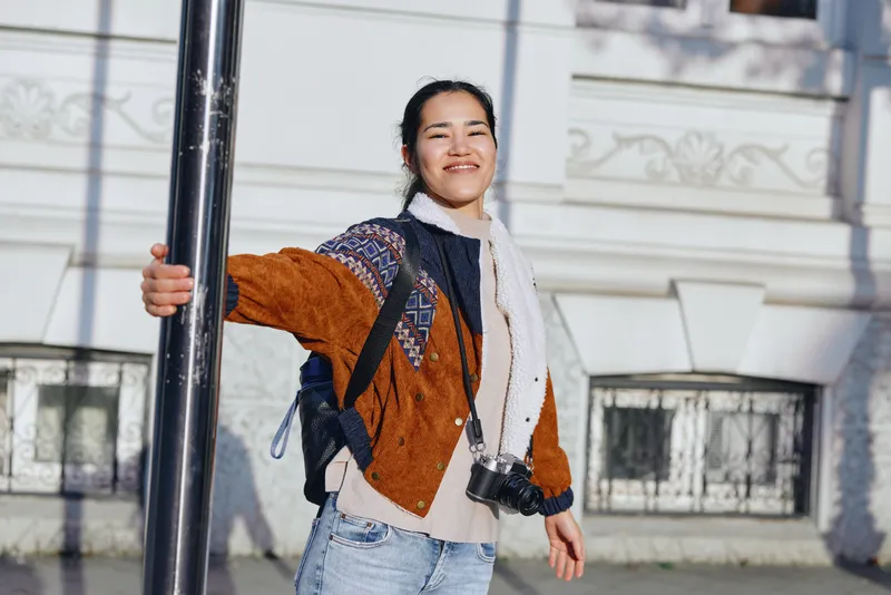 Candid outdoor street fashion shot of a smiling woman with a backpack leaning on a pole, wearing a warm jacket and scarf in bright urban light