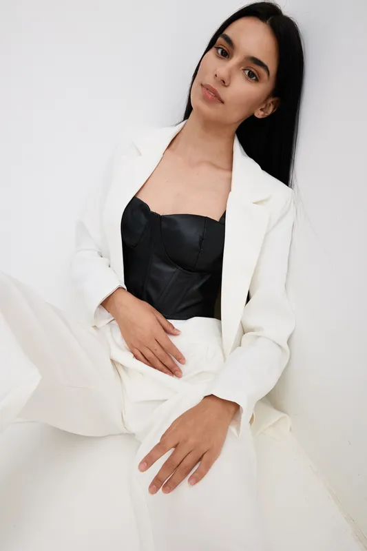 elegant woman wearing white suit and black corset posing against blank white wall, featuring long black hair and natural makeup in minimalistic style