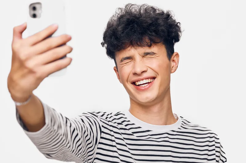 Young man smiling happily with eyes closed taking a selfie wearing a striped shirt against a white background, displaying joy and relaxed mood.