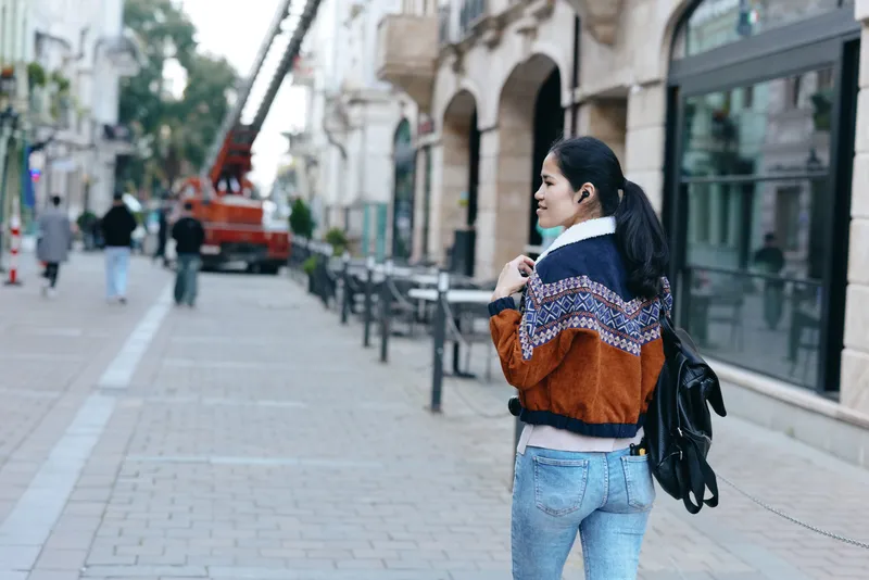 Woman with backpack walks along a city sidewalk wearing a patterned jacket and jeans, glancing back as urban street scene unfolds with buildings and pedestrians.