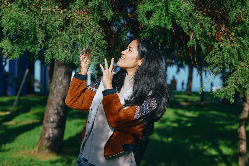 Nature scene featuring a woman beneath lush trees, fingers brushing wet branches as droplets fall in an outdoor park, stylish jacket, serene candid moment.
