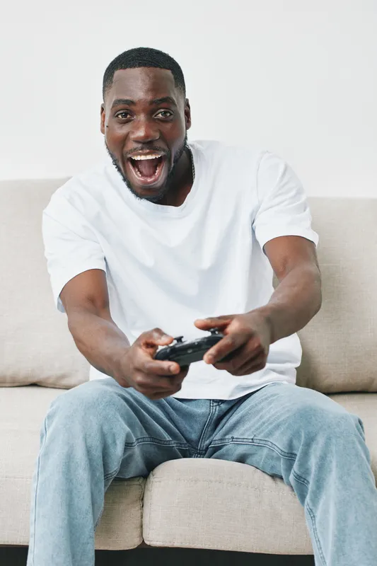 Excited young Black man playing video games at home, wearing a white t shirt and denim jeans, conveying joy and enthusiasm in a bright living room
