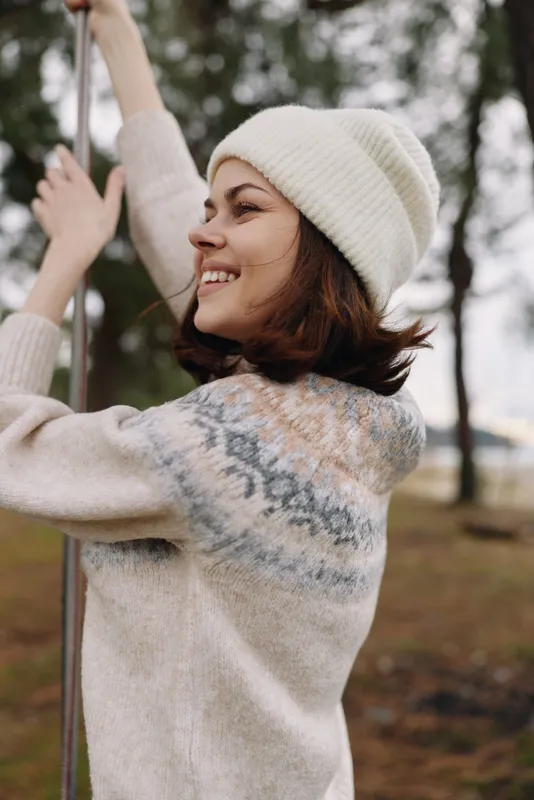 Woman wearing a knit sweater and beanie outdoors in a park, smiling while gripping a pole during winter, showcasing casual leisure and seasonal fashion.