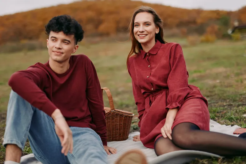 Candid outdoor picnic portrait of a young couple sitting on a blanket with a wicker basket, sharing a relaxed, authentic and genuine moment that conveys credible everyday connection.