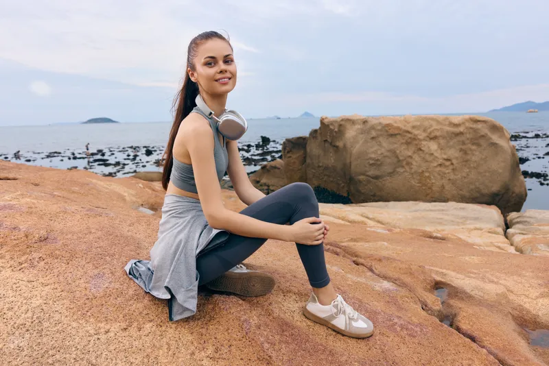 Young woman in activewear sitting on rocky beach, smiling with headphones, enjoying healthy lifestyle at seaside, ideal for wellness and fitness themes