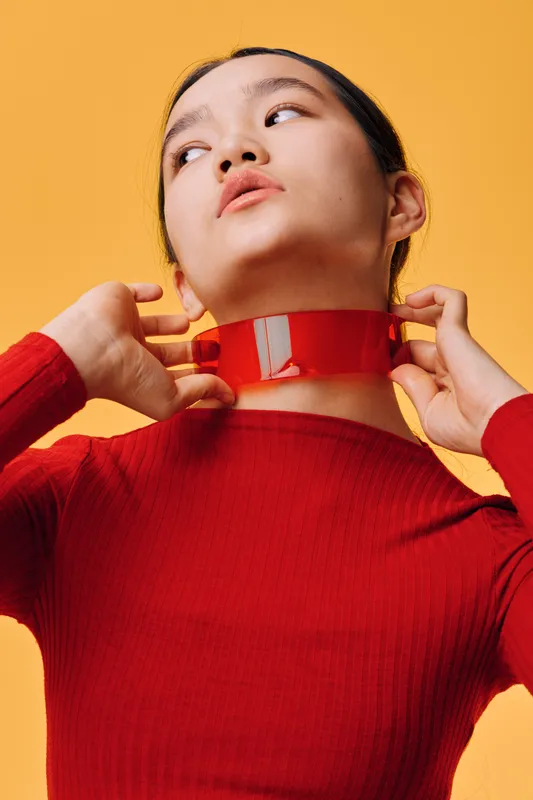 Woman in red sweater adjusting shiny red vinyl collar on neck with hands, looking away on bright yellow background, fashion accessory and modern style portrait in studio.