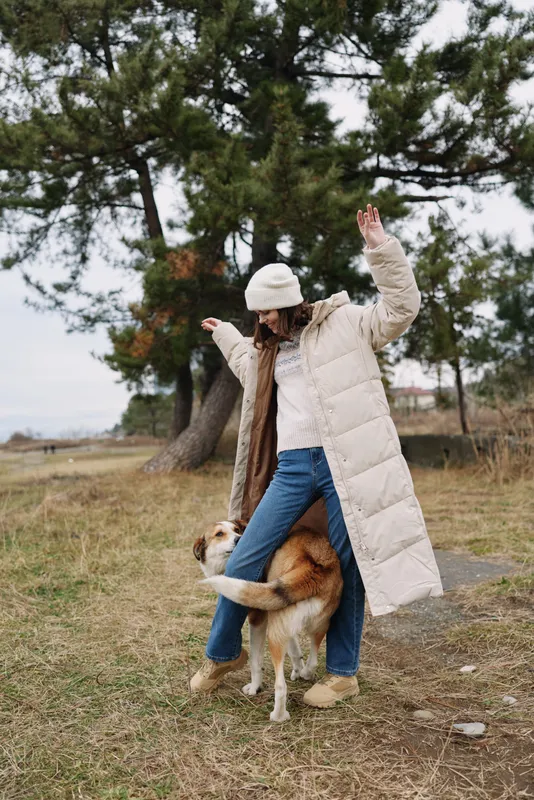 Woman wearing a long beige winter coat and beanie dances with a playful dog in an open outdoor field among trees, capturing joyful motion and natural style.