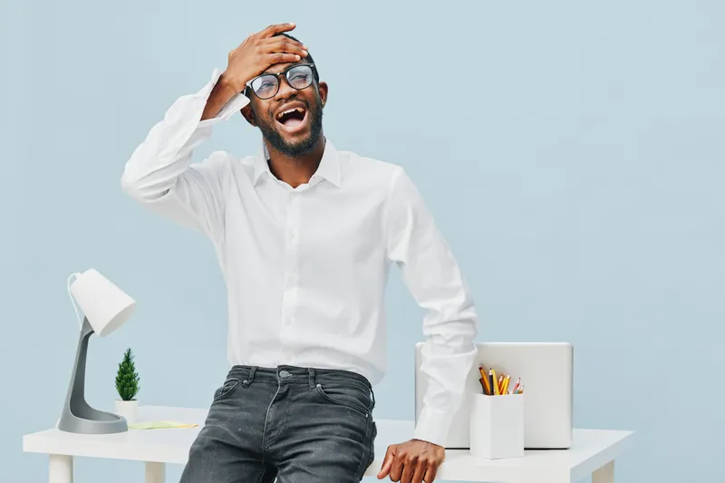 Smiling happy young african american man wearing glasses and white shirt sitting at modern office desk with laptop, plant, and stationery isolated on light blue background. Office lifestyle concept, c