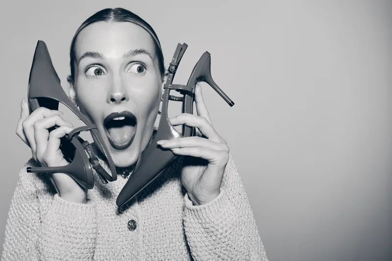 Surprised Caucasian woman with fair skin holds red high heels near face, wearing knitted sweater, isolated on grey background.