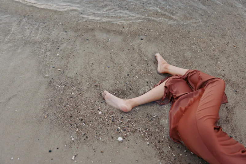 A lone person lies on wet sand near the shoreline, legs extended in rust-colored pants, toes pointed, as gentle waves lap the beach and leave subtle trails.