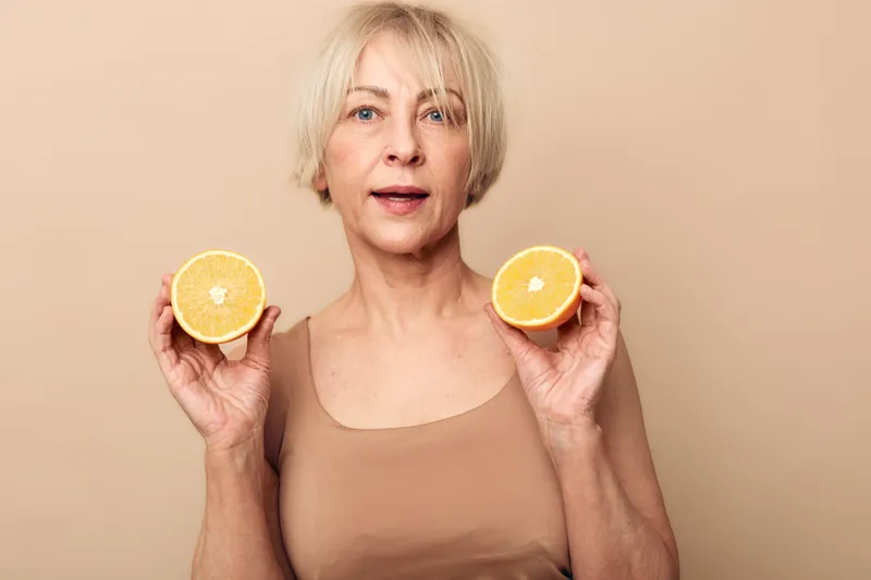 Mature woman with short blonde hair holds two halves of fresh orange fruit against beige background. Concept for healthy lifestyle, nutrition, wellness, and natural vitamin sources.
