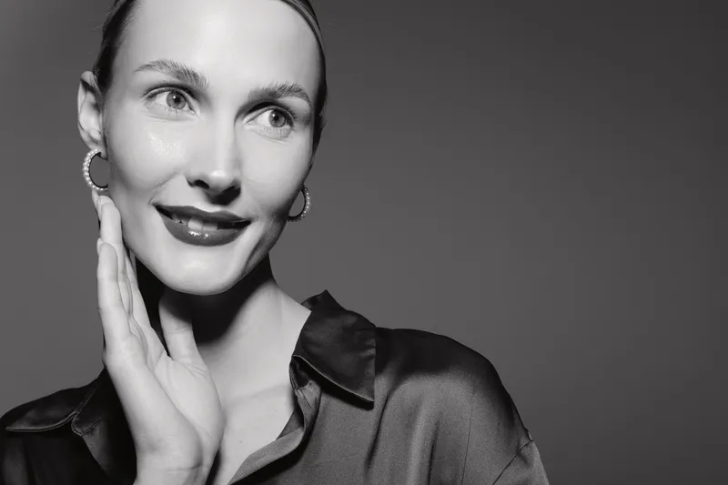 Young Caucasian woman with fair skin smiles gently, touching her face with hand. Studio portrait in black and white with soft lighting and smooth satin blouse.
