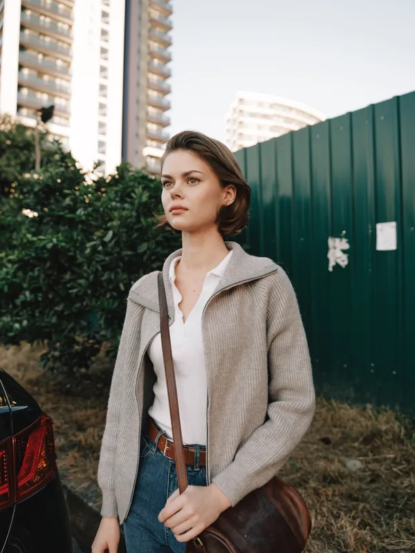A poised professional woman stands outdoors in an urban setting, wearing a white blouse and light cardigan with a crossbody bag, projecting a calm, focused, and approachable business mood.