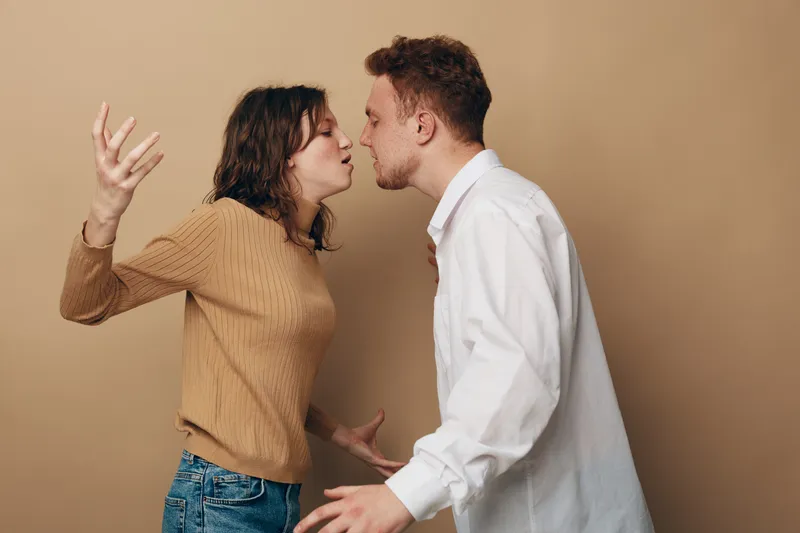 Angry young couple arguing indoors, woman and man facing each other with tense expressions. Concept of relationship conflict, disagreement, and emotional tension in casual clothing.