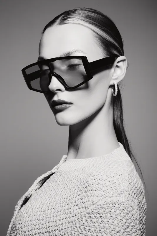 Elegant Caucasian woman with pale skin wearing large geometric sunglasses and textured white sweater. Studio portrait with dramatic lighting and minimalist style.