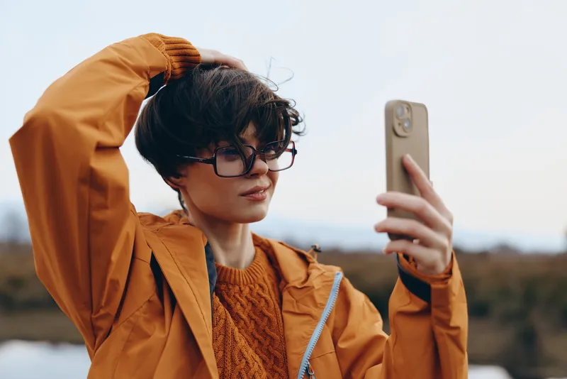 Woman lifestyle selfie outdoors, orange jacket woman holds phone for portrait, glasses and confident gaze, wind moved hair, sunny background, casual fashion, relaxed mood