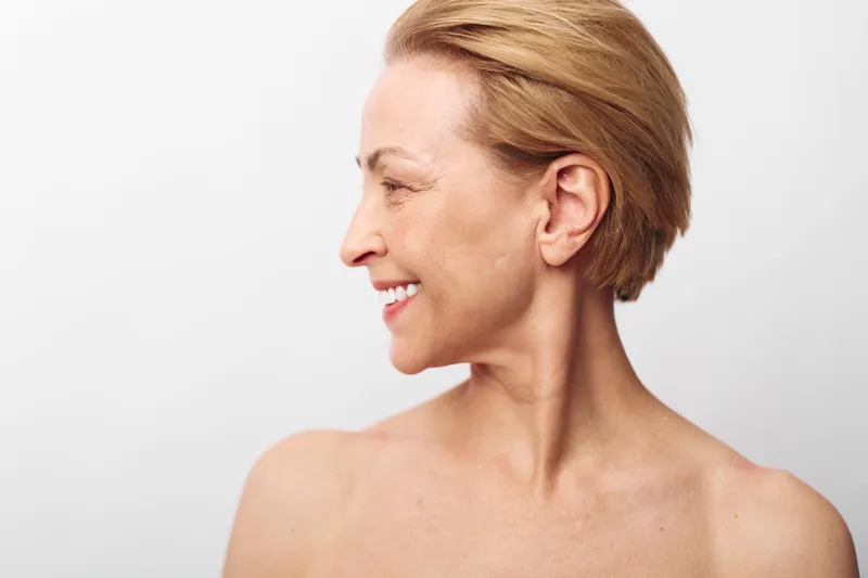 Smiling middle aged woman with short blonde hair looks sideways, bare shoulders visible. Studio portrait on white background. Concept of happiness, natural beauty, healthy skin and aging gracefully.