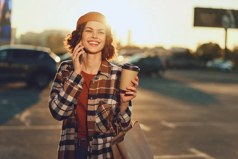 Woman smiling on phone holding coffee and shopping bag in urban street at golden hour, candid lifestyle portrait with authenticity, mindful living and emotional storytelling vibe.