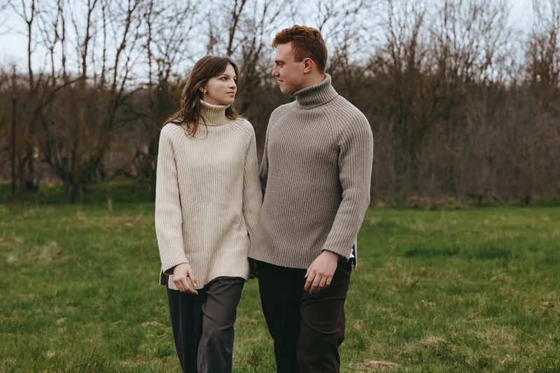 A couple in chunky knit sweaters share a quiet moment in a grassy park, holding hands and looking at each other. A warm lifestyle portrait capturing connection, comfort, and everyday romance in an