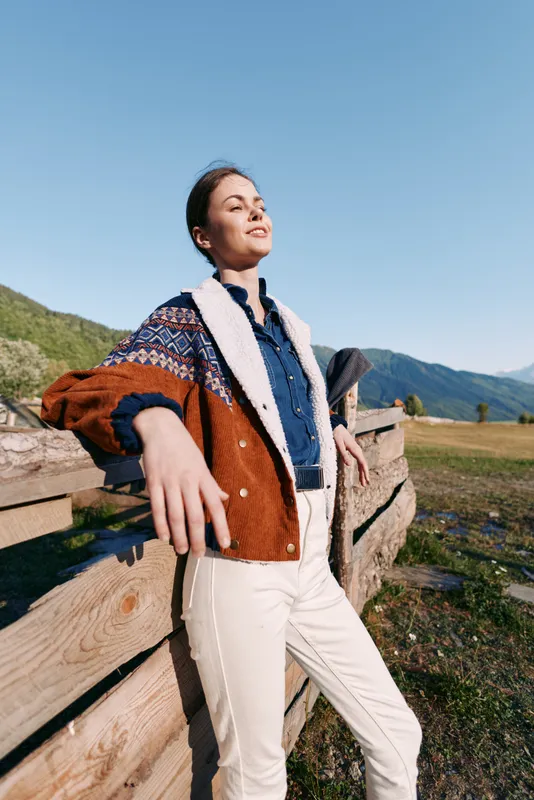 Woman leaning on wooden fence in countryside with mountains and blue sky, with a smile and relaxed pose. Fashionable jacket and casual outfit in a confident outdoor portrait and nature scenery.