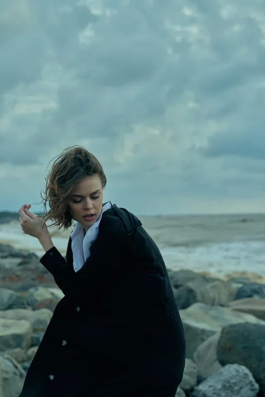 Woman on a rocky coast wearing a dark coat, wind in hair, contemplative mood by the sea, fashionable outdoor scene showing resilience and style against a stormy sky and crashing waves