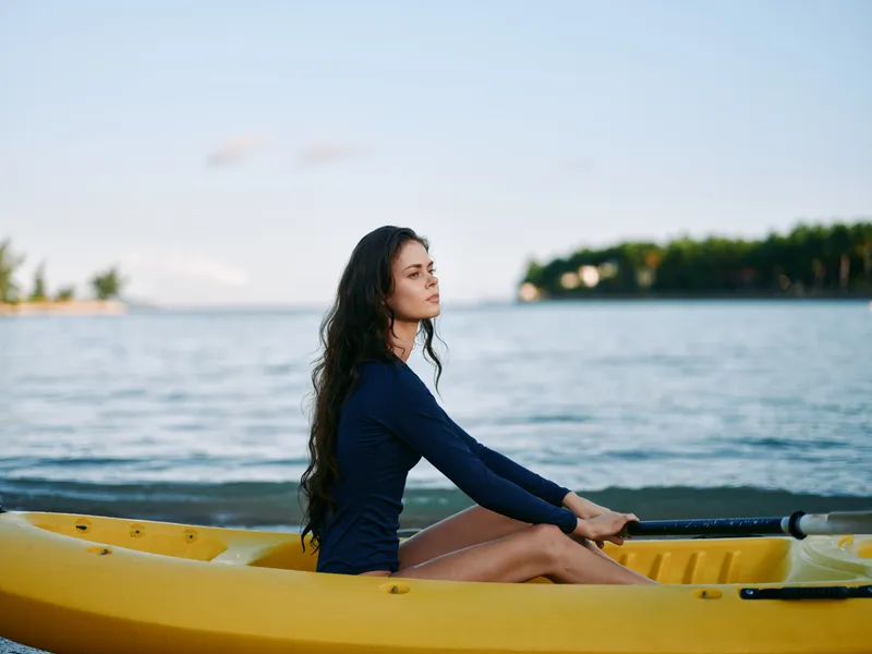 A lone woman sits in a yellow kayak on calm water, facing left, wearing a navy blue long-sleeve outfit, with flowing curly hair, bathed in soft sunset light.