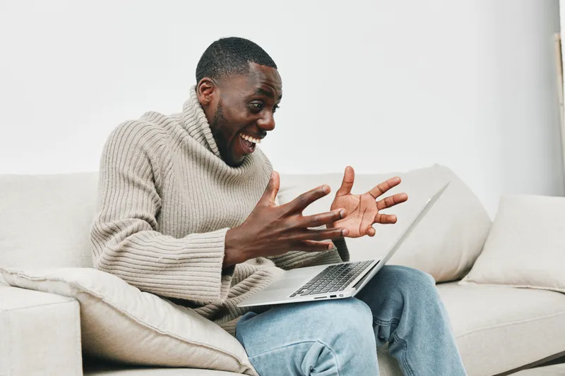 Excited man in cozy sweater celebrating success while using laptop at home, showcasing joy and positive emotions in a bright, modern living space