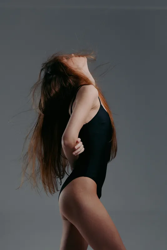 Graceful woman dancer posing with long hair flip, wearing black bodysuit against gray background, expressive motion and elegance.