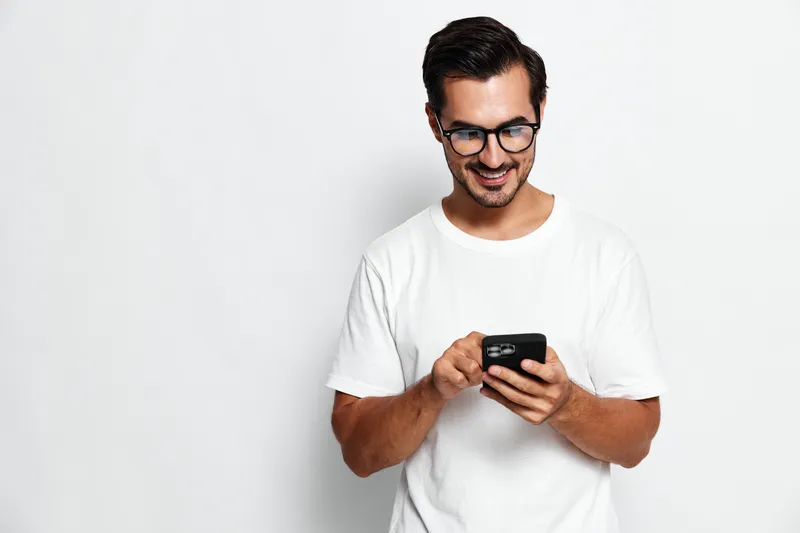 Smiling young man wearing glasses and a white t-shirt, happily interacting with his smartphone, standing against a solid light background. People lifestyle concept.
