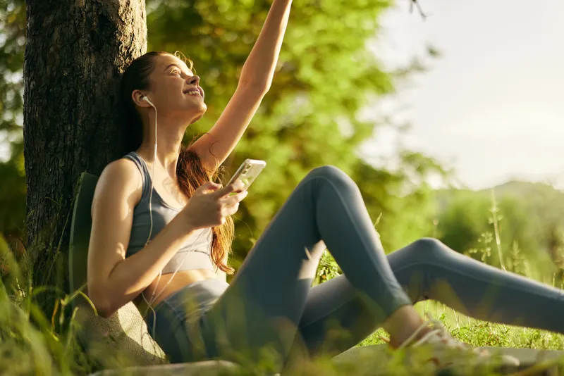 Young woman enjoying music in nature with smartphone, wearing sporty outfit, relaxed expression under sunlight, summertime vibe and fresh greenery