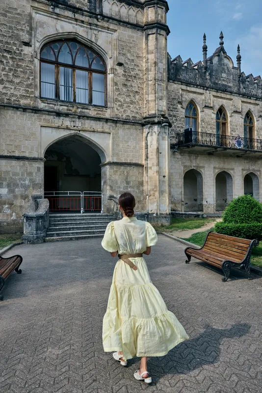 A woman in a pale yellow vintage dress walks toward a grand historic castle facade, creating a graceful scene of timeless elegance and exploration