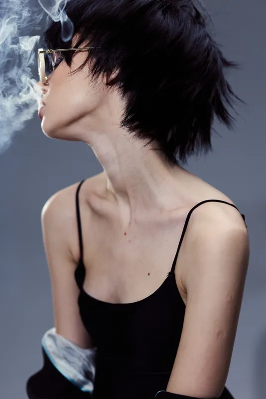 Young woman with short hair blowing smoke, wearing black sleeveless top and sunglasses, stylish and modern portrait
