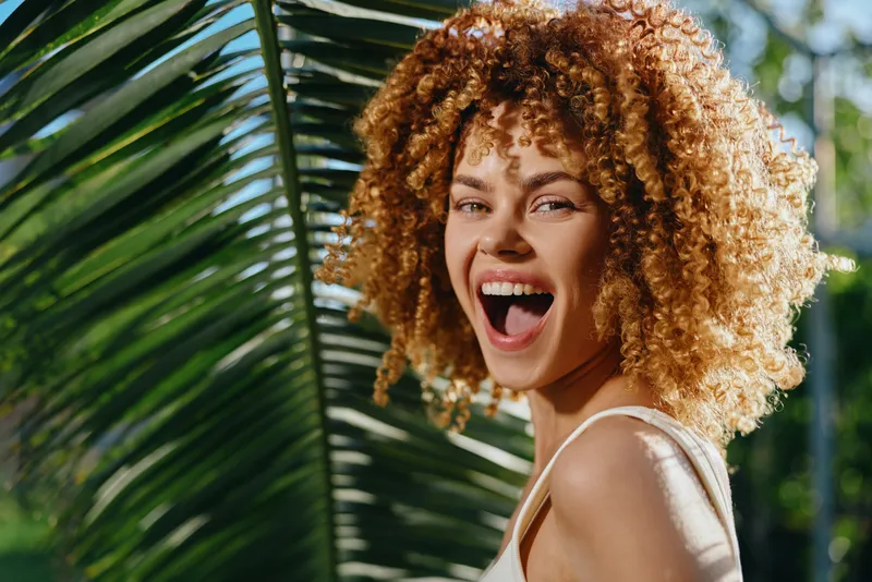 Smiling woman with curly hair enjoying sunny tropical outdoors, natural light highlighting her joyful expression and green palm leaves background.