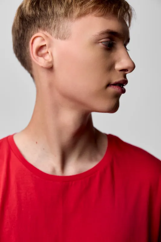 Profile view of a young boy in a red shirt, calm expression, clean studio lighting, neutral backdrop, focus on facial features and casual fashion.