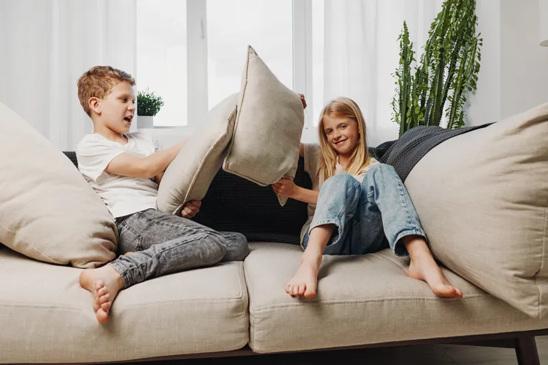 Two happy children having pillow fight on beige sofa in bright living room. Casual clothing, playful mood, natural light, cozy interior with plants and soft cushions. Family fun concept.