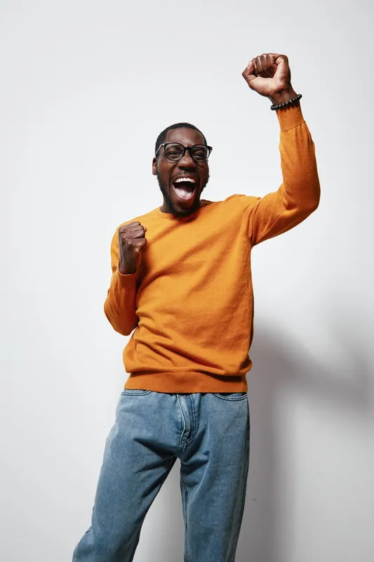 African American man with dark skin wearing glasses and orange sweater celebrating with raised fists and joyful expression against white background in studio.