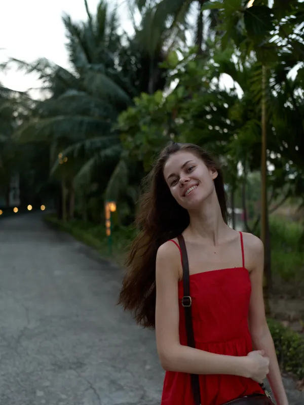 smiling young woman in red dress standing outdoors on a pathway surrounded by lush green trees and palm leaves, enjoying a peaceful moment in nature