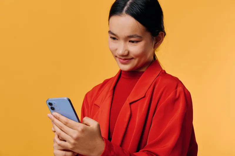 Woman in red jacket smiling and looking at smartphone screen with interest on yellow background. Young female holding mobile device with both hands for texting or browsing.