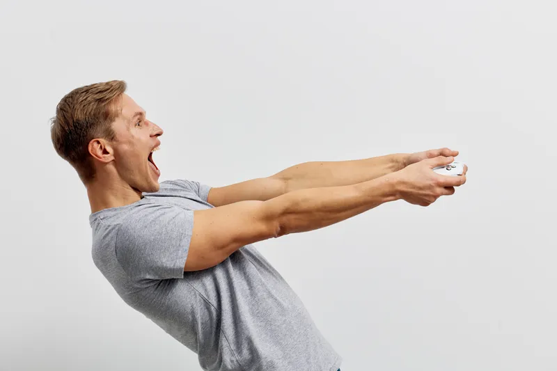 Young smiling man in casual grey t-shirt stretching his arms forward while holding a TV remote control, isolated on white background. Studio shot demonstrating excitement and enthusiasm. People lifest