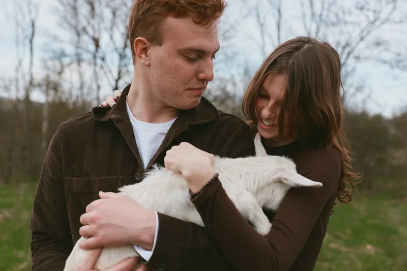Joyful couple outdoors with a white goat kid, warm embrace, casual clothing, soft natural light, rural setting, candid moment, genuine connection, peaceful mood, rustic charm