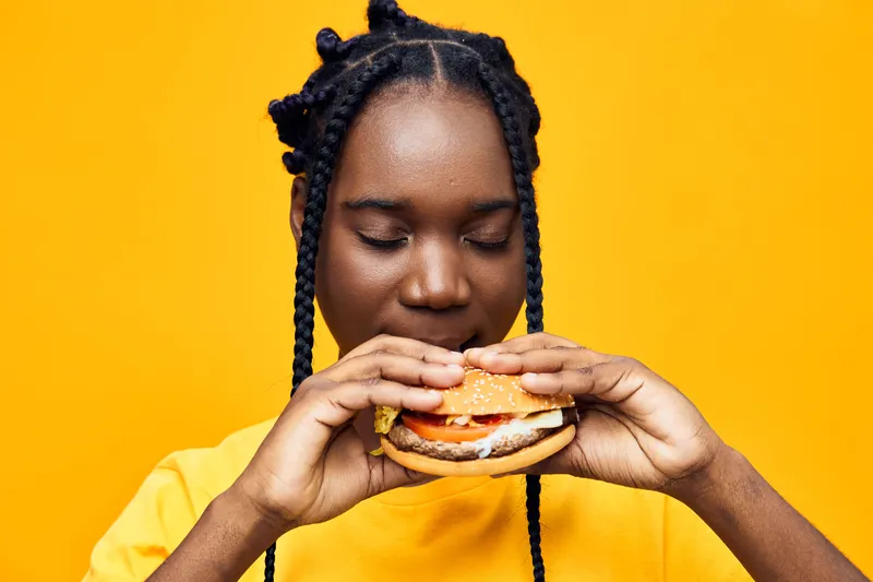 Young Black woman enjoying a delicious, colorful burger against a vibrant yellow background, capturing the joy of healthy eating and vibrant lifestyles