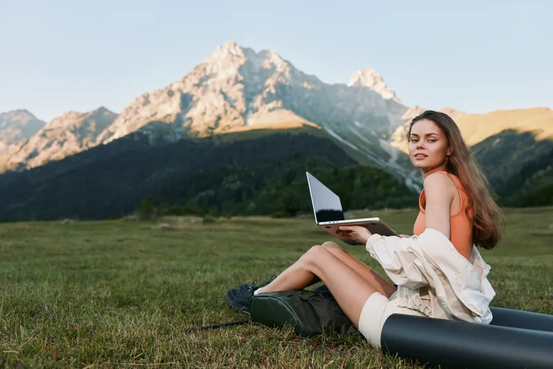 A cheerful young woman sits on grass with a laptop outdoors beside scenic mountains, smiling while typing, creating a relaxed productive vibe in a natural workspace.