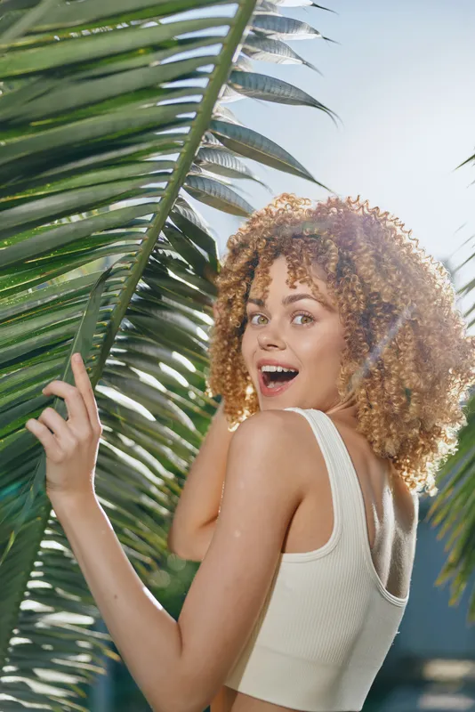 smiling woman with curly hair standing behind tropical palm leaves outdoors in bright sunlight, wearing a white sleeveless top, summer vibe