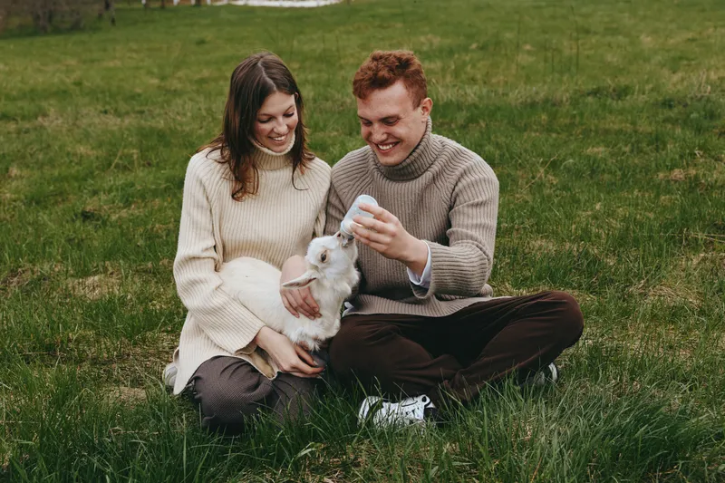 A young couple sits on the grass feeding a small white baby goat with a bottle while enjoying a relaxed lifestyle moment outdoors in casual autumn clothing.